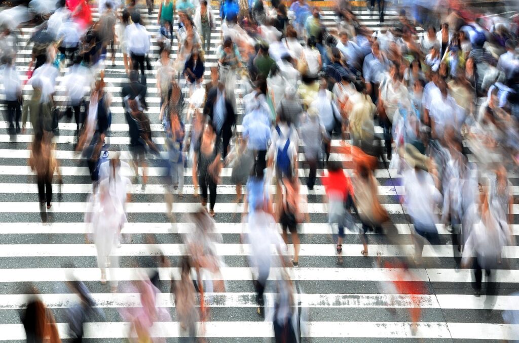We zien een enorm zebrapad met heel veel overstekende mensen in beide richtingen. De foto is genomen met een lange sluitertijd waardoor de mensen vervagen tot onherkenbare schimmen wat extra accent geeft aan de dynamiek in de foto.