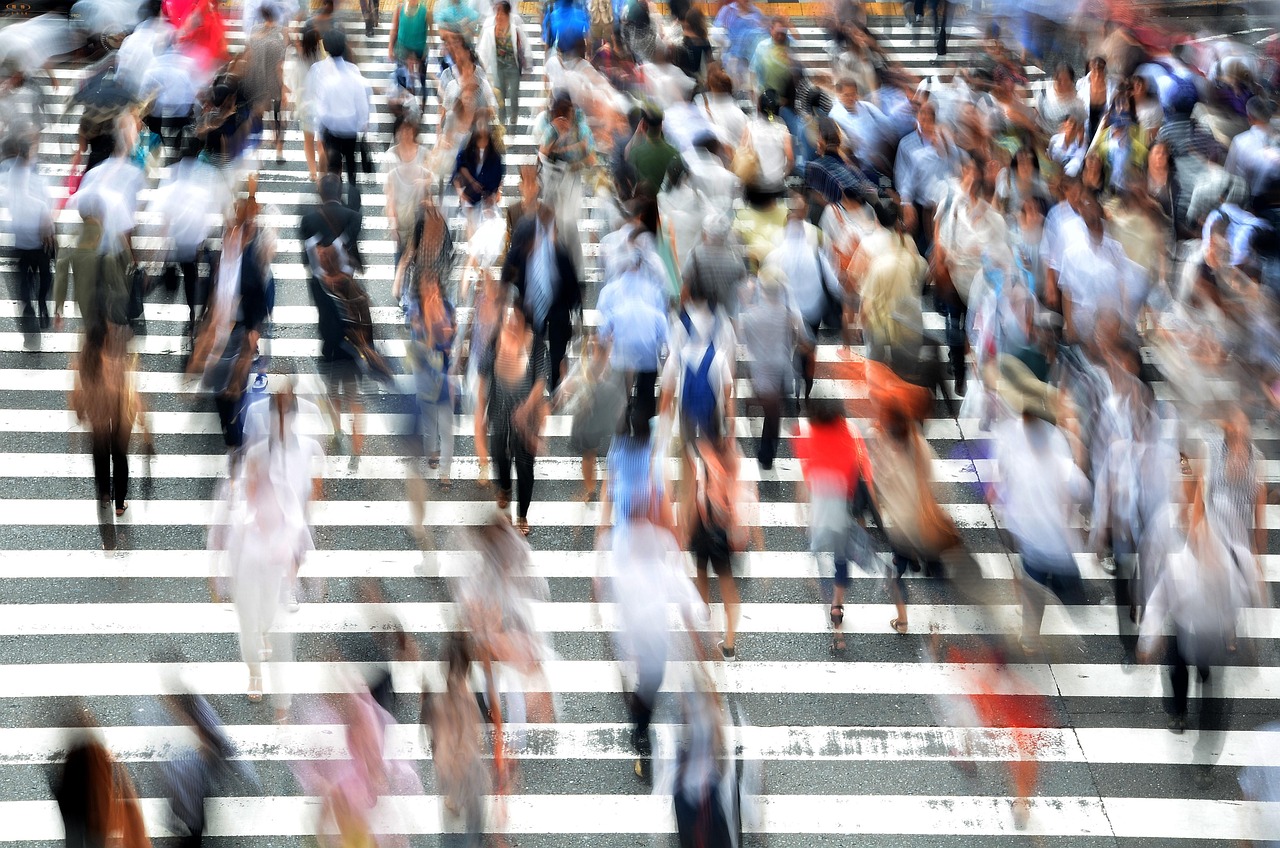 We zien een enorm zebrapad met heel veel overstekende mensen in beide richtingen. De foto is genomen met een lange sluitertijd waardoor de mensen vervagen tot onherkenbare schimmen wat extra accent geeft aan de dynamiek in de foto.