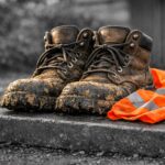 Een paar modderige, versleten werkschoenen staan op een donkergrijze, licht gevlekte stoeprand met werksporen. Achter de schoenen ligt een helder oranje veiligheidsvestje met reflecterende strepen, nonchalant op de grond. De achtergrond is zacht zwart‑wit en filmisch vervaagd, waardoor de schoenen en het vestje in kleur het menselijke verhaal van arbeid en vermoeidheid benadrukken.