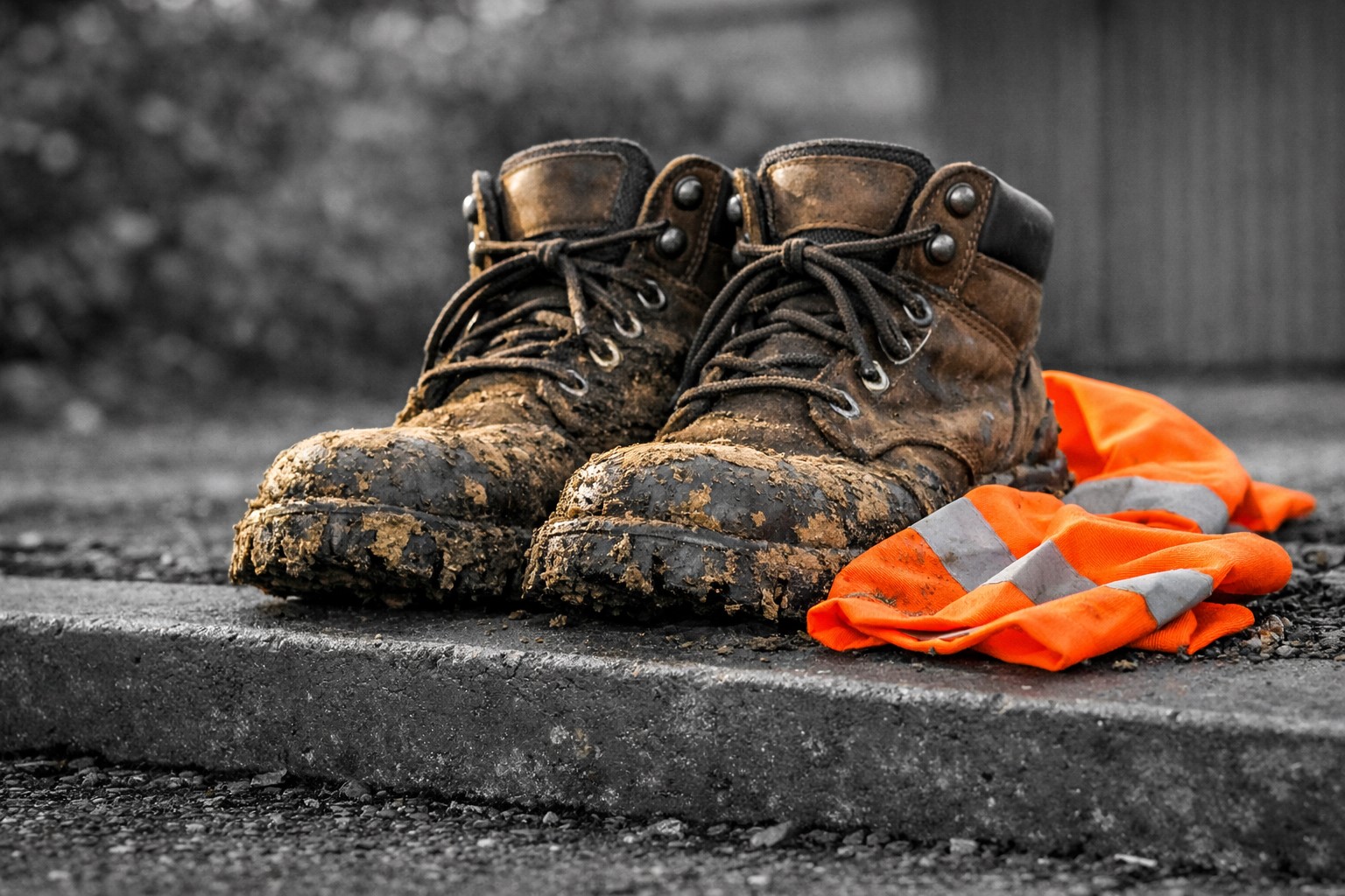 Een paar modderige, versleten werkschoenen staan op een donkergrijze, licht gevlekte stoeprand met werksporen. Achter de schoenen ligt een helder oranje veiligheidsvestje met reflecterende strepen, nonchalant op de grond. De achtergrond is zacht zwart‑wit en filmisch vervaagd, waardoor de schoenen en het vestje in kleur het menselijke verhaal van arbeid en vermoeidheid benadrukken.
